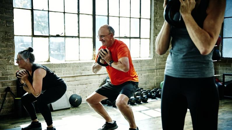 Atletas idosos e sorridentes fazendo agachamentos com kettlebell durante a aula de fitness - Idosos treinando, atividade física exercício idosos