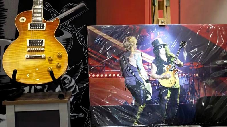 Exposição mostra guitarra elétrica amarela estilo sunburst em pedestal. Atrás, fotografia grande de dois músicos em palco iluminado; um deles toca guitarra semelhante, enquanto o outro usa instrumento diferente. Luzes fortes e fundo vermelho compõem a cena.