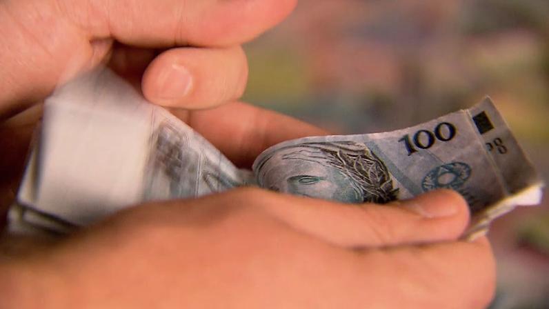 Mãos seguram e contam notas de 100 reais, moeda brasileira. Cédulas azuis exibem número “100” e parte da efígie da República.
