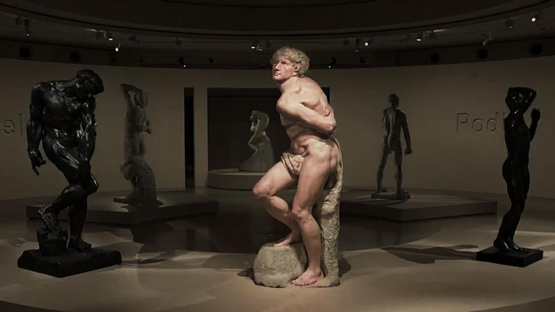 Exposição em galeria ou museu com esculturas sobre plataformas circulares. Obras representam figuras humanas em diferentes poses, algumas em bronze escuro e outras em mármore claro. No centro, grande escultura de homem musculoso parcialmente saindo de bloco de pedra. Ao fundo, inscrições nas paredes remetem a nomes de escultores famosos. Iluminação de teto destaca texturas e detalhes das peças.