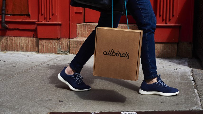 A person wearing Allbird shoes carrying an Allbirds box walking down a city sidewalk