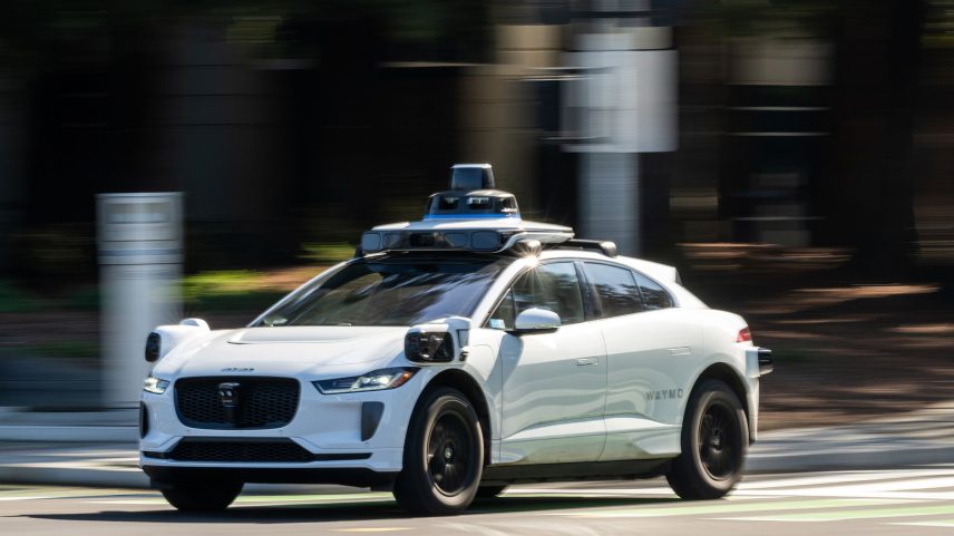 A Waymo autonomous taxi drives through a city intersection.