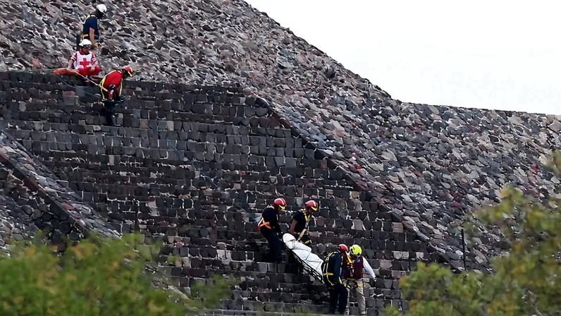 A body is carried on a stretcher down the steps of a grey stone pyramid.