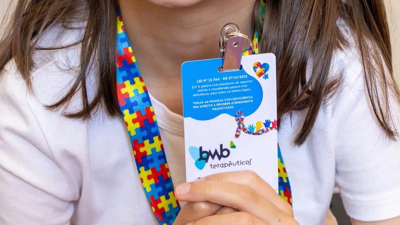 Pessoa veste camisa branca e segura crachá preso a cordão colorido com peças de quebra-cabeça em vermelho, azul, amarelo e verde. No crachá há texto em português mencionando lei sobre direitos do autismo e prioridade de atendimento para pessoas com deficiência.