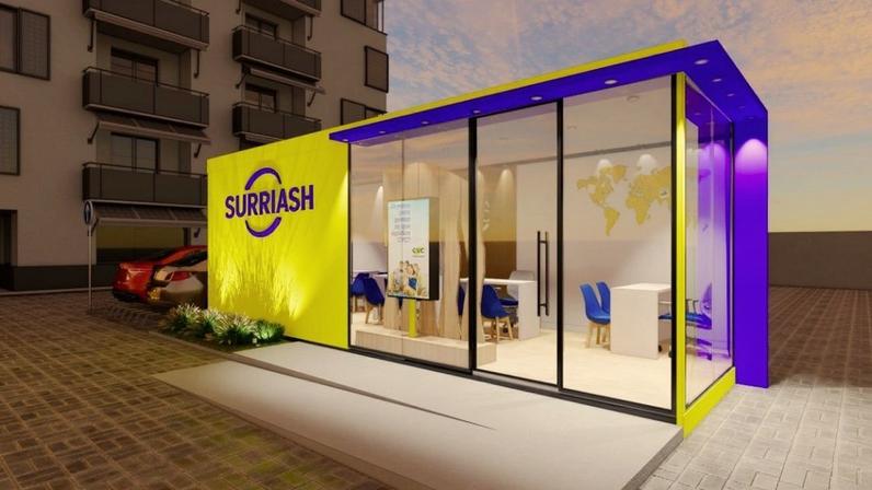 Quiosque modular CVC amarelo e roxo com janelas de vidro, em área externa pavimentada com edifício e carro.