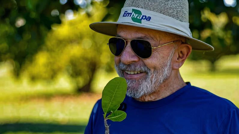 Lula usando uma camiseta azul e um chapéu da Embrapa segura uma muda de planta
