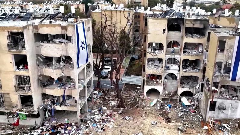 Dois prédios residenciais aparecem com grandes danos estruturais, paredes e varandas destruídas. No chão, há destroços espalhados. Nas fachadas, pendem bandeiras de Israel.
