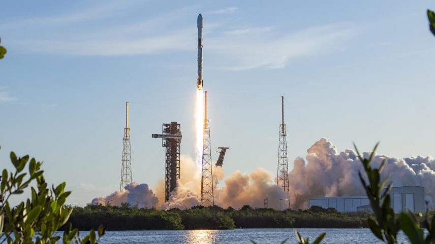 Image shows a rocket launching vertically in a morning blue sky with white clouds at Cape Canaveral Space Force Station in Florida on Saturday, April 11, 2026. The launch reflects in a nearby body of water and shrubs.
