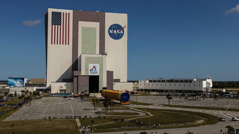 The image shows NASA's Kennedy Space Center iconic Vehicle Assembly Building with an American flag painted on the building along with the NASA and Artemis insignias in a blue sky with some clouds on Tuesday, April 28, 2026. People gathered to watch the transport of the Artemis III core stage, an orange yellow that will launch during the Artemis III mission. White buildings, grass, and a parking lot can also be seen in the photo.