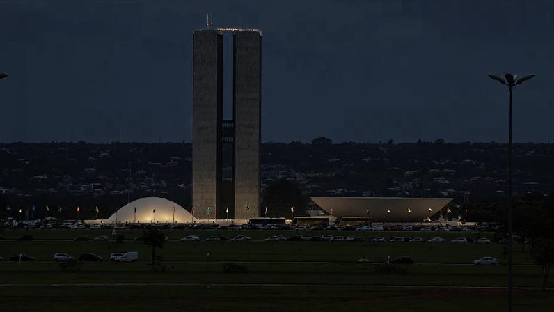 Vista do Congresso Nacional em Brasília, com as duas torres retangulares ao centro, as cúpulas branca e côncava dos plenários à frente, gramado amplo em primeiro plano e carros circulando pela via