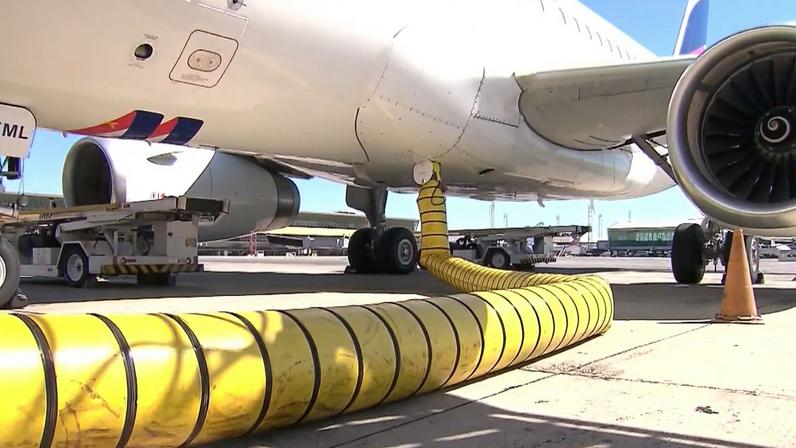 Avião estacionado no pátio de um aeroporto durante abastecimento, com mangueira amarela conectada à parte inferior da aeronave e equipamentos de serviço ao redor
