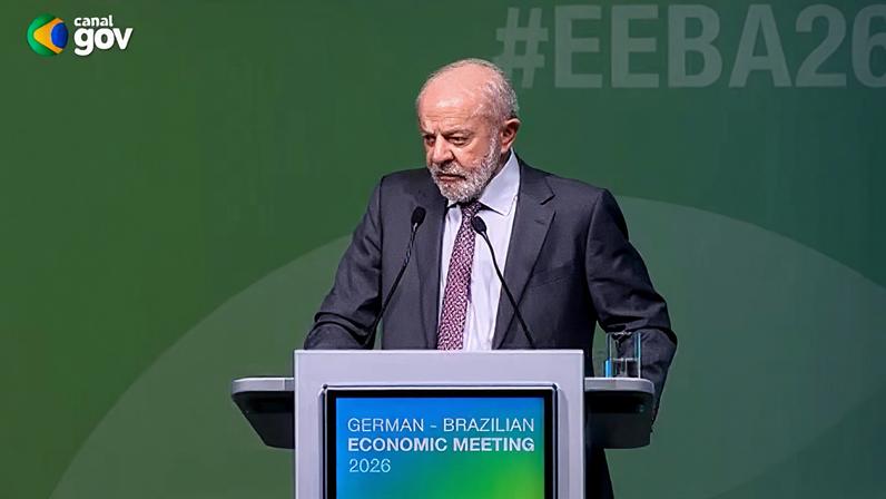 Lula veste terno e está em pé atrás de um púlpito, falando em um evento oficial. O púlpito tem uma tela com o texto “German‑Brazilian Economic Meeting 2026”