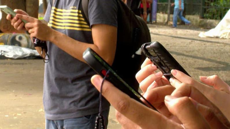 Pessoas em pé ao ar livre segurando e usando celulares, com foco nas mãos e nos aparelhos
