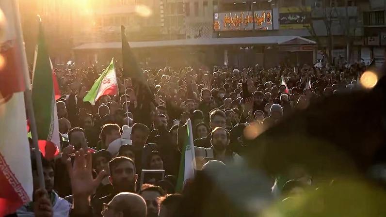 Grande multidão está reunida em uma área urbana ao ar livre durante o pôr do sol. Muitas pessoas seguram bandeiras iranianas. A luz intensa do sol atravessa a cena por trás, criando um brilho forte na parte superior da imagem. Ao fundo, é possível ver prédios, letreiros e estruturas da cidade.