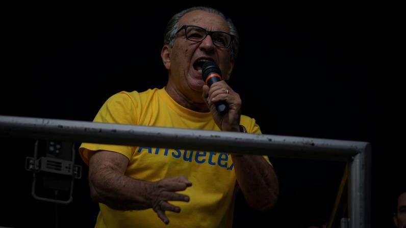Imagens coloridas mostram homem de meia idade, calvo, vestindo camisa amarela, com microfone na mão esquerda, em cima de um carro de som