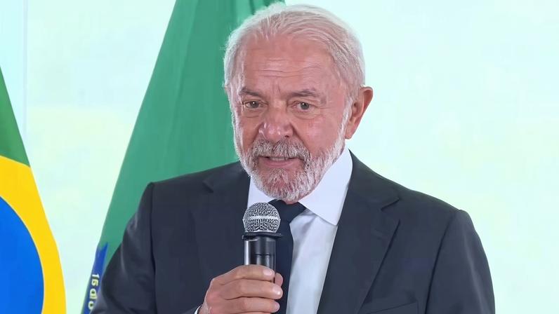 Homem de cabelos brancos e barba usa terno escuro, camisa branca e gravata, falando em microfone. Atrás dele aparecem duas bandeiras, uma do Brasil e outra verde.