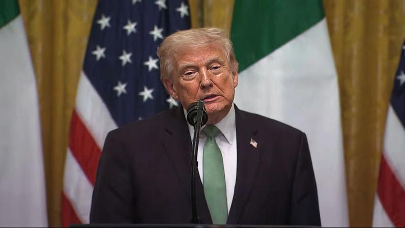 Donald Trump usando terno escuro, camisa branca e gravata verde, posicionado atrás de um púlpito com microfone, com bandeiras dos Estados Unidos ao fundo