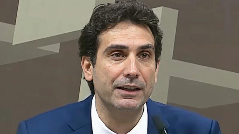 Pessoa veste terno azul com camisa branca e gravata azul, falando em microfone. Fundo apresenta formas geométricas em tons de bege e marrom, sugerindo ambiente formal como conferência, reunião ou entrevista.