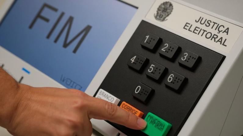 Close up da mão de uma pessoa apertando o botão "confirma" na urna eletrônica.