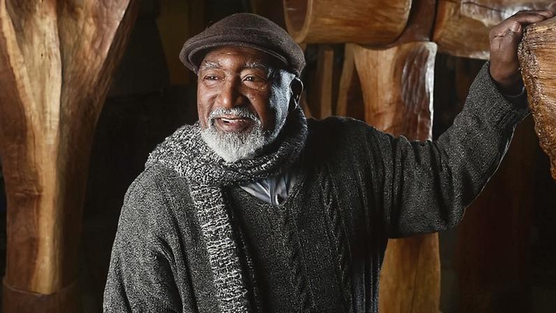 Portrait of Thaddeus Mosley wearing a scarf with one hand on a carved wooden sculpture.