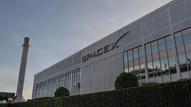 Fachada da SpaceX, com letras cinzas em um prédio baixo branco, céu está cheio de nuvens