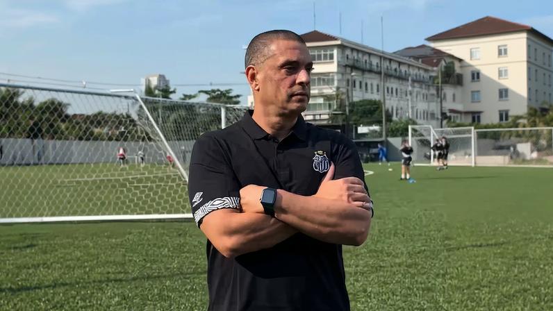 Pessoa em campo de futebol, de braços cruzados, veste camisa preta com logotipo do Santos FC e símbolo da Umbro na manga. Ao fundo, traves de gol e pessoas participando de atividades esportivas.