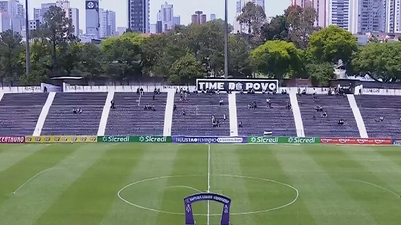Estádio de futebol com arquibancadas parcialmente ocupadas. No topo, faixa grande traz a frase “TIME DO POVO”. Gramado bem cuidado mostra linhas brancas do círculo central. Estrutura roxa em arco está posicionada no meio do campo. Placas de publicidade exibem marcas conhecidas.