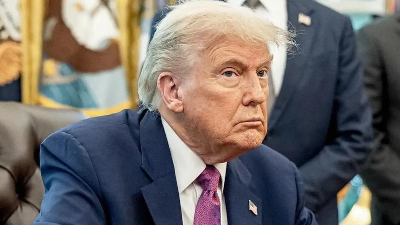 Donald Trump, de terno azul com pin da bandeira, sentado em um escritório. Ele tem um olhar sério e pensativo.