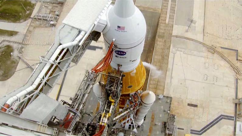 A imagem mostra uma visão aérea de um foguete espacial de grande porte posicionado em uma plataforma de lançamento. O foguete tem corpo branco com uma seção inferior em tom laranja e está conectado a várias estruturas metálicas de suporte, braços de serviço e tubulações. Cabos grossos e mangueiras aparecem ligados à lateral do veículo, indicando procedimentos técnicos de preparação. A base ao redor é composta por concreto claro, trilhos e marcas no solo que delimitam a área operacional. O enquadramento destaca a complexidade da infraestrutura de lançamento, com passarelas, torres e sistemas de acesso distribuídos ao redor do foguete, em um ambiente industrial amplo e organizado.