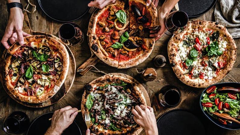 Família ou amigos em uma festa de pizza. Vista de cima de pessoas pegando e comendo diversos tipos de pizza e bebendo vinho tinto sobre uma mesa rústica de madeira.