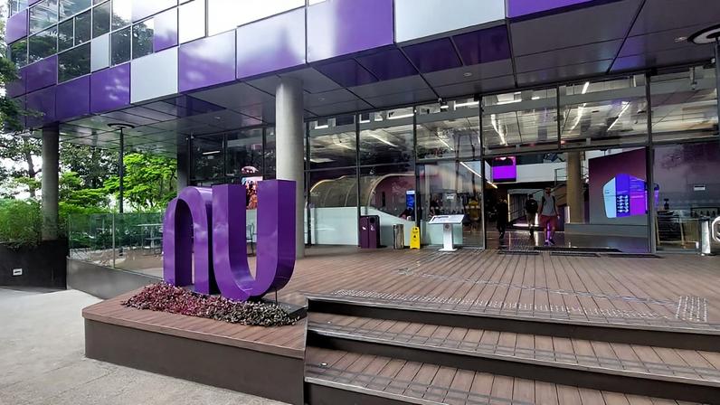 Logo roxo 'nu' do Nubank na entrada de um prédio moderno. Fachada roxa e cinza, com deck de madeira