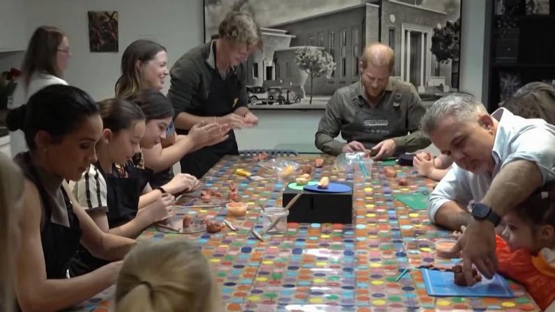 Grupo de pessoas reunidas em torno de mesa coberta por toalha colorida de bolinhas. Adultos e crianças participam de oficina, moldando peças de argila com ferramentas e recipientes pequenos.