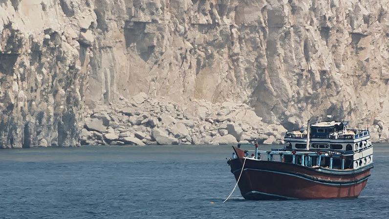 Um barco está ancorado no meio do Estreito de Ormuz. Atrás dele é possível ver um grande paredão de pedra.