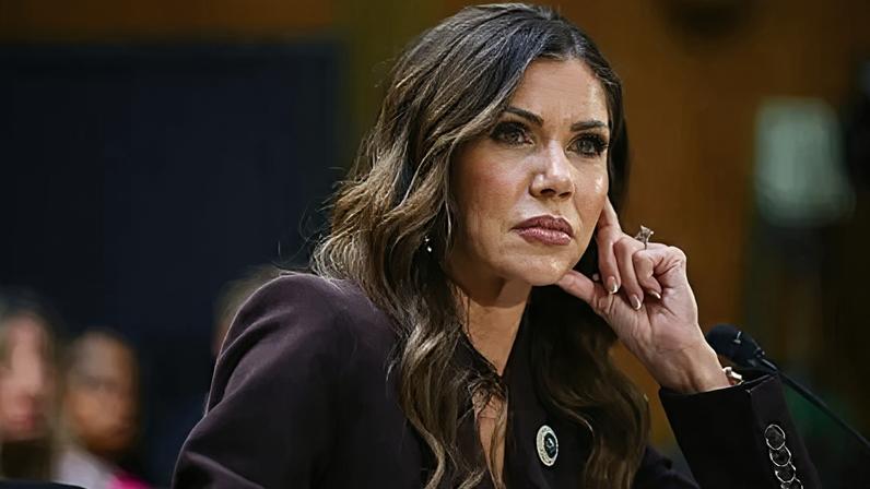 Kristi Noem tilts her head to the right as she props her head up with her left hand. Her hair falls along the side of her face and she is wearing an eggplant coloured blazer with a broach on the left lapel.