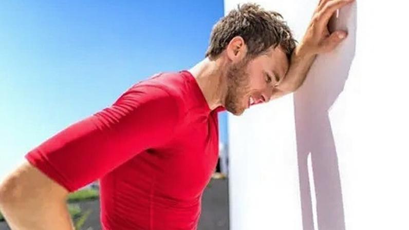 Getty Images A sweating man with brown hair and wearing a red top leans against a white wall in the sun because he is overheating.