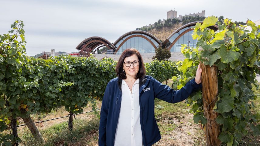 Marilena Bonilla standing between two rows of vines, with her hand up against a wooden post and the winery in the background