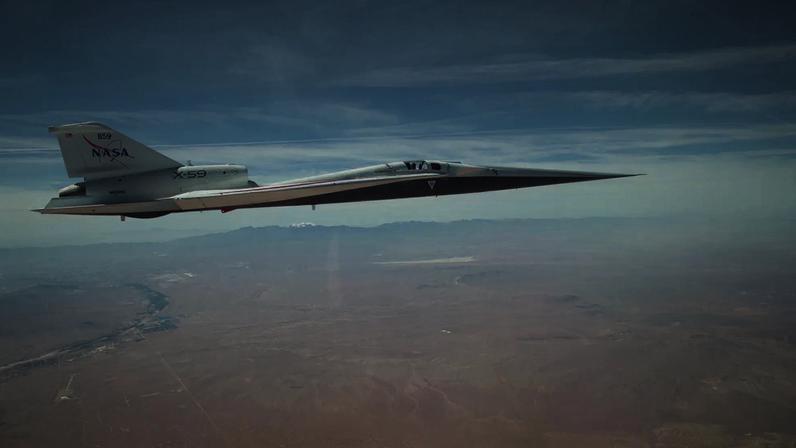 NASA’s X-59 quiet supersonic research aircraft flies above the Mojave Desert on a clear day. The aircraft is white with light gray, red, and blue accent colors. A NASA logo is visible on its tail, along with the number 859 above it. It appears to be flying level over the desert land-scape, and in the distance, you can see a mountain range on the horizon.