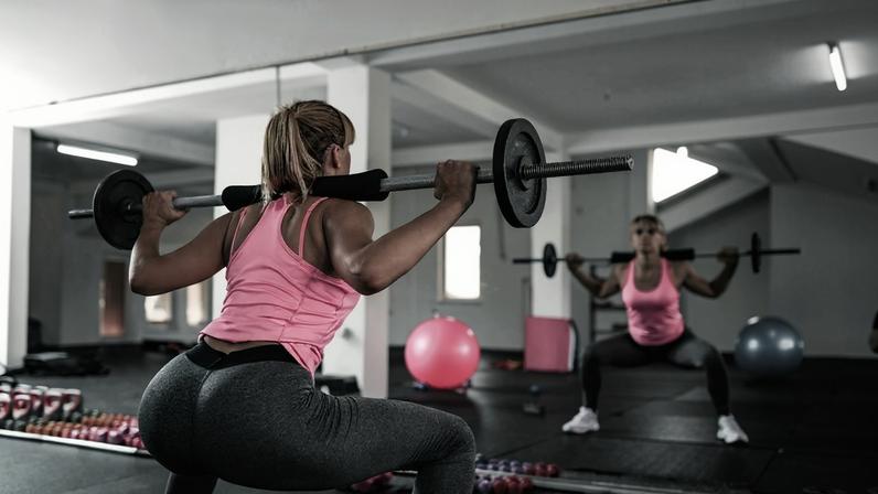 Mulher fazendo agachamentos com pesos na academia. Metrópoles