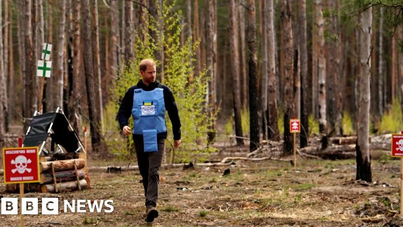 Prince Harry, a white man with short red hair, walking through a woodland in a blue vest and grey trousers. He is walking past signs that have a skull and bones image on them and writing.
