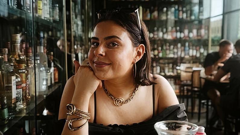 Abi A young woman in a bar with lots of bottles of drink in cabinets behind her. You can see the top half of her body. She is wearing a black top and resting her chin on her hand. She has sunglasses pinning back her hair and has golden earings, necklace and bracelet.