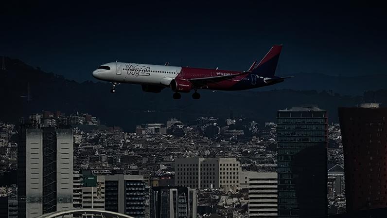 Getty Images A white, pink and blue airplane with "Wizz" written on the side flies low over a city, with wooded hills in the background.