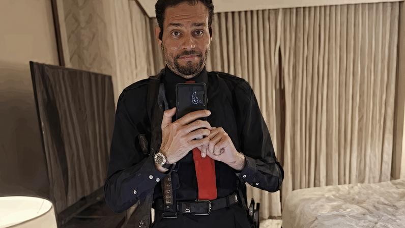Homem aparece vestido de preto, com gravata vermelha e carrega uma faca, um coldre de ombro para arma e uma bolsa para munição