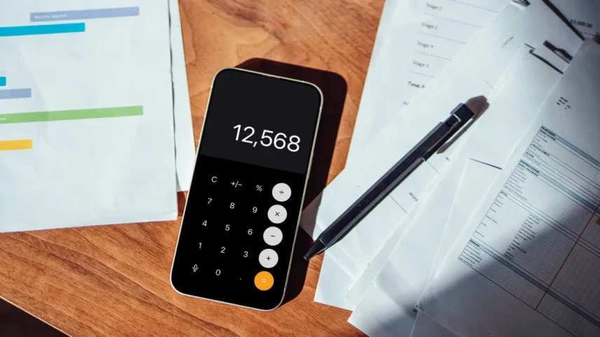 Ambiente de escritório com mesa de madeira, documentos e smartphone. Smartphone mostra tela de calculadora com número 12.568, cercado por papéis, gráficos coloridos e caneta preta.