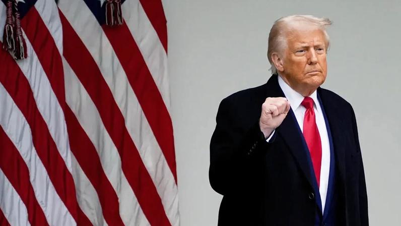 Donald Trump, punho cerrado, de casaco escuro e gravata vermelha, com bandeiras americanas ao lado.