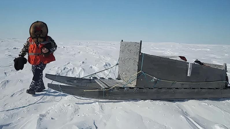A man walking on snow with a fur collar and hi-vis jacket walks by a komatik, an indigenous sled.