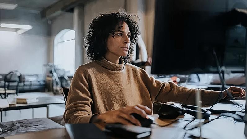 Getty Images Woman with dark curly hair, wearing tan polo neck jumper, works on a computer in an office