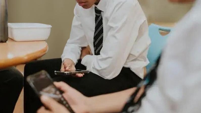 Getty Images Two students in school uniform sit indoors during a break, looking down at smartphones held in their hands.