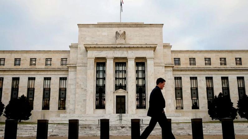 Homem de terno caminhando em frente ao edifício do Federal Reserve, com bandeira e selo da águia, sob céu nublado.