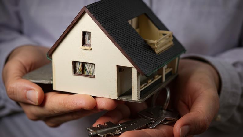 Foto de uma miniatura de uma casa segurada na palma de duas mãos, ao lado de uma chave, ilustrando o "Minha Casa, Minha Vida"