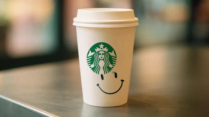 Copo descartável de papel com tampa branca e logotipo verde da Starbucks, destacando uma sereia de duas caudas no centro. Abaixo, um desenho de rosto sorridente. Fundo desfocado de cafeteria e superfície metálica refletem luz.